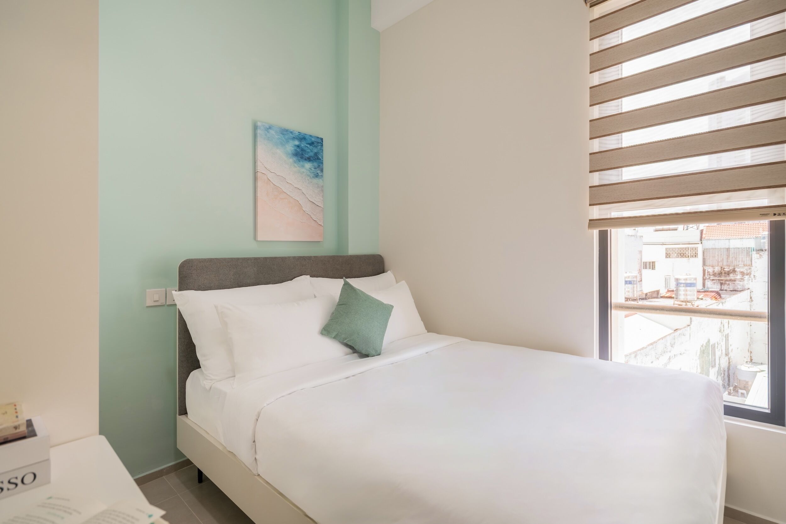 Een netjes opgemaakt bed met wit beddengoed en een groen kussen, een muur met een schilderij, en een raam met zonwering.