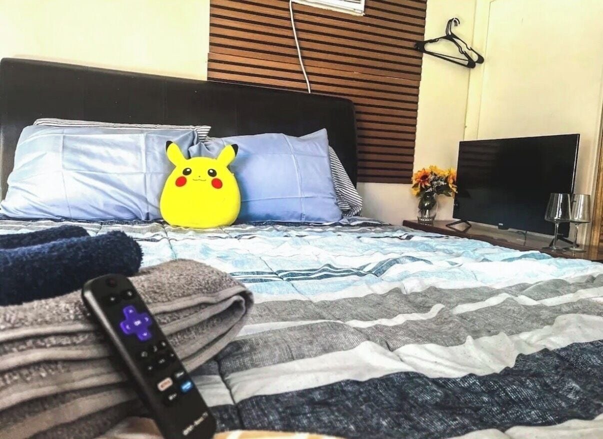 Ein Bett mit einem gemusterten Überwurf, Kissen, einer Fernbedienung, einem Fernseher und einer wandmontierten Klimaanlage.