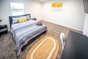 A bedroom with a bed, a desk, a chair, a rug, a nightstand, and a window.