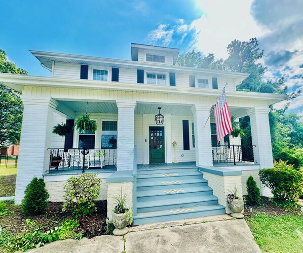 A two-story white house with a front porch, a green door, and a flag.