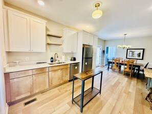 Una cocina moderna con armarios blancos, un refrigerador de acero inoxidable y un comedor con sillas y mesa de madera.