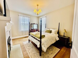 Un dormitorio con cama con dosel, mesita de noche, lámpara, cómoda, espejo y ventana con persianas.