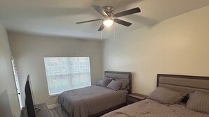 Ein Schlafzimmer mit zwei Betten, einem Deckenventilator, einem Fernseher auf einem Holztisch und einem Fenster mit Jalousien.