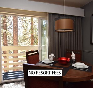 Un área de comedor con una mesa dispuesta para dos, un tazón rojo, platos blancos y una copa de vino. Un letrero dice "NO RESORT FEES".