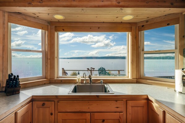 A kitchen with a large window overlooking a lake, wooden cabinets, and a stainless steel sink.
