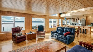 A spacious living area with wooden floors and walls, large windows offering views of the water, a kitchen with modern appliances, and leather furniture.