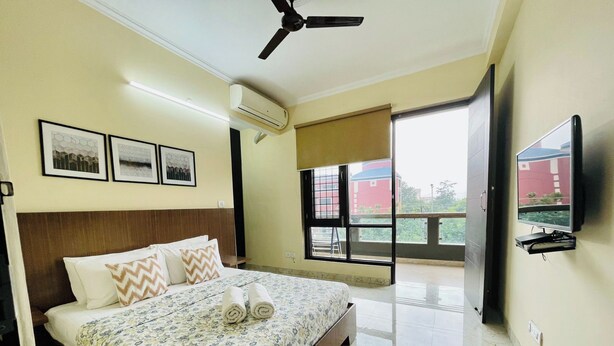 A hotel room with a bed, ceiling fan, air conditioning unit, framed pictures, a TV, and a balcony with a view. - Jolo Hotel Gurgaon and Birthday Venues (Gurugram)
