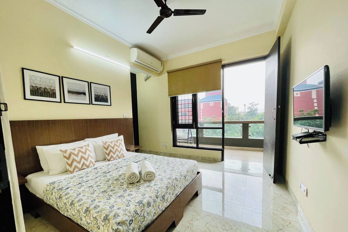 A hotel room with a bed, ceiling fan, air conditioning unit, framed pictures, a TV, and a balcony with a view.