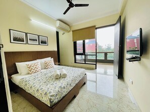 A hotel room with a bed, ceiling fan, air conditioning unit, framed pictures, a TV, and a balcony with a view. - Jolo Hotel Gurgaon and Birthday Venues (Gurugram)