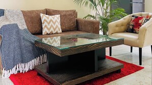 A living room with a brown sofa, a glass coffee table, a red rug, and a beige sofa. There are three framed pictures on the wall and a plant in the corner. - Jolo Hotel Gurgaon and Birthday Venues (Gurugram)