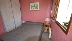 Een kleine kamer met een enkel bed, houten frame, nachtkastje met lamp, klein plankje met bloemen en een schilderij aan de muur.