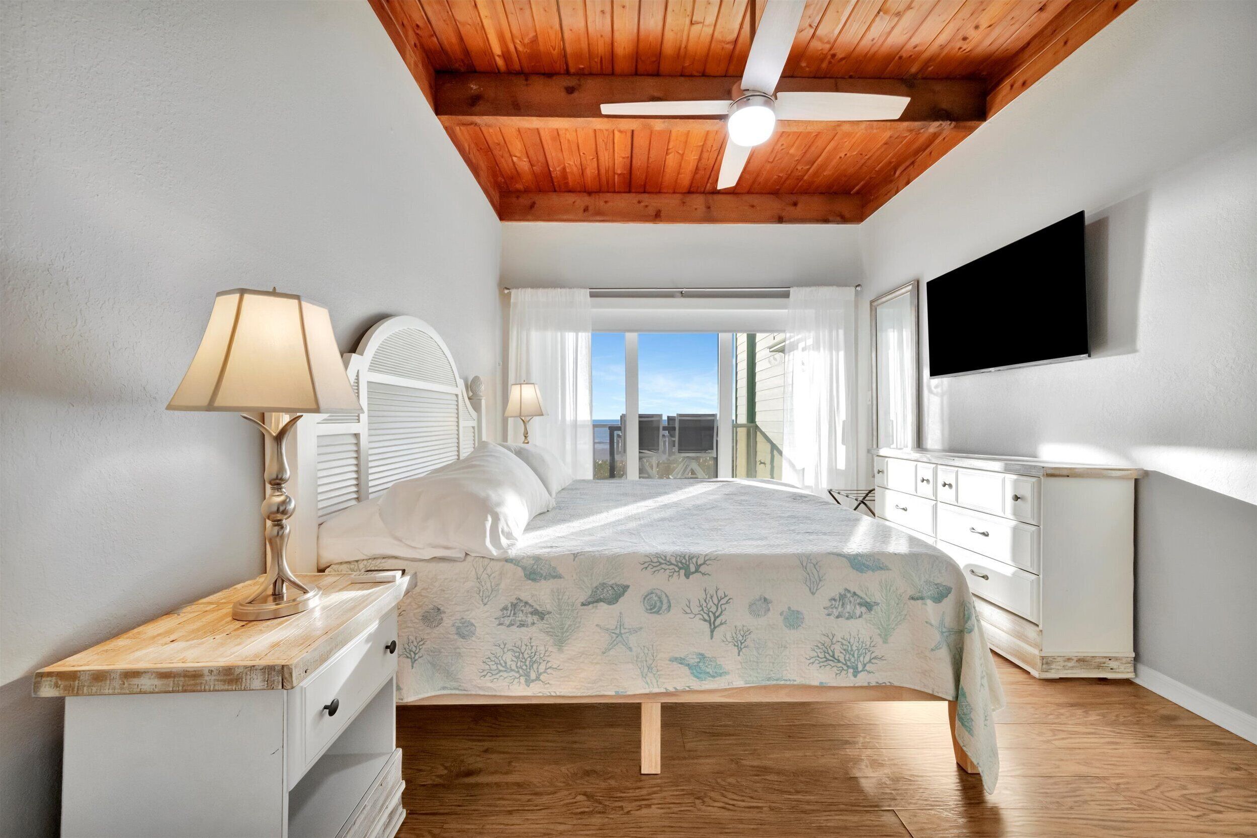 A bedroom with a wooden ceiling, a bed with a blue ocean-themed bedspread, a bedside table with a lamp, a dresser, and a TV mounted on the wall.