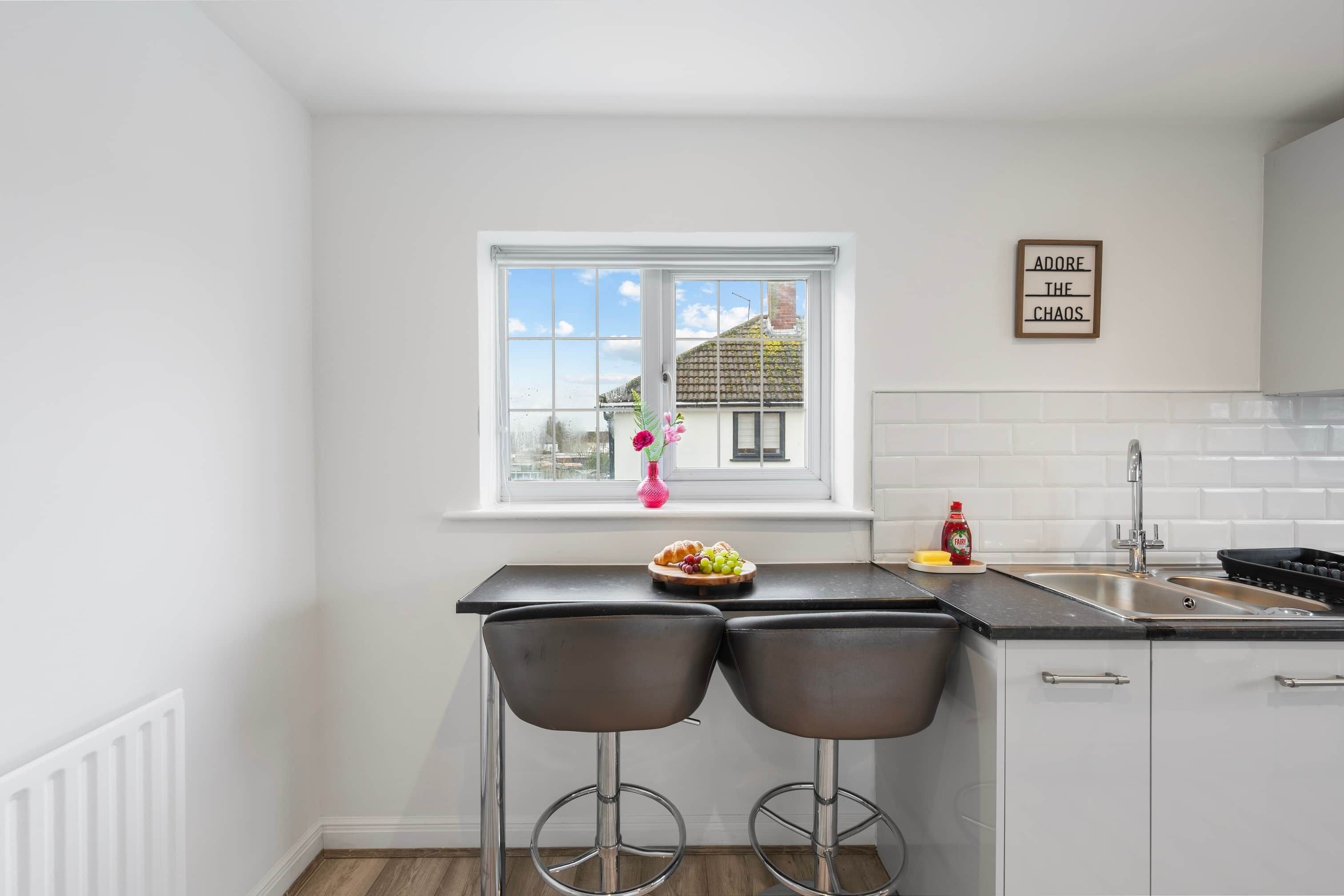 Een keuken met een raam, twee barkrukken, een fruitschaal, een spoelbak en een bordje aan de muur.