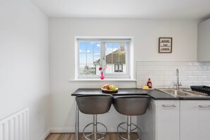 A kitchen with a window, two bar stools, a fruit bowl, a sink, and a wall-mounted sign. - Cosy Welcome Stay (Slough)