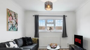 En moderne stue med en sort sofa, et rundt sofabord, et fjernsyn og et stort vindue med gardiner.