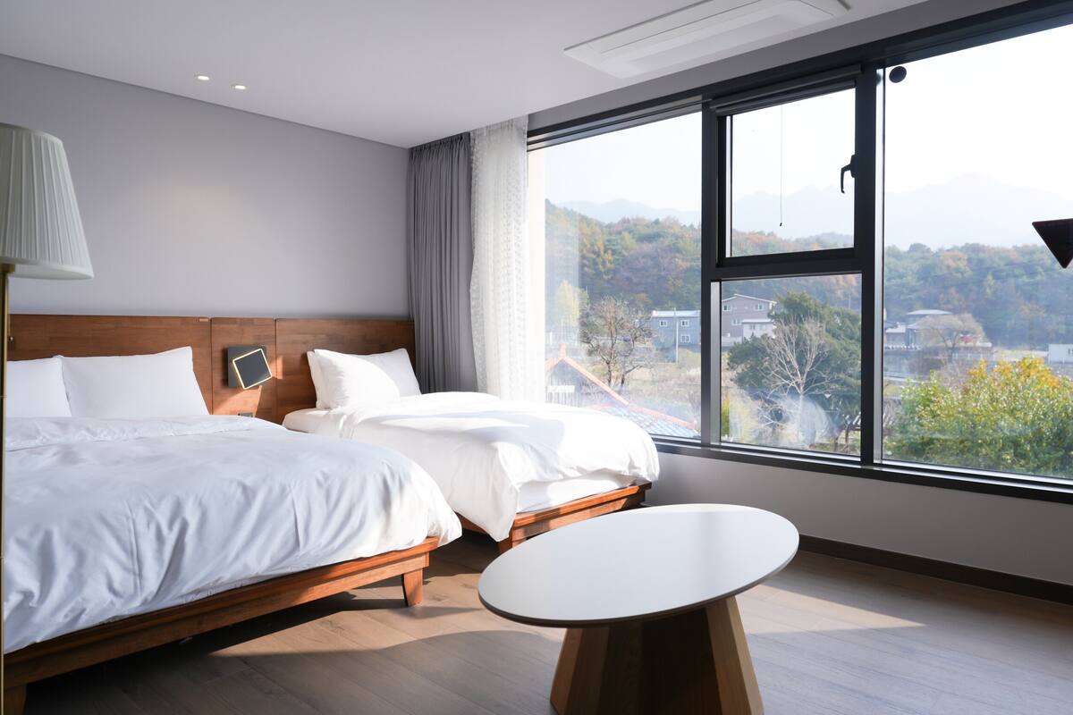 A modern hotel room with two beds, a bedside table, and a large window offering a view of the city and mountains.