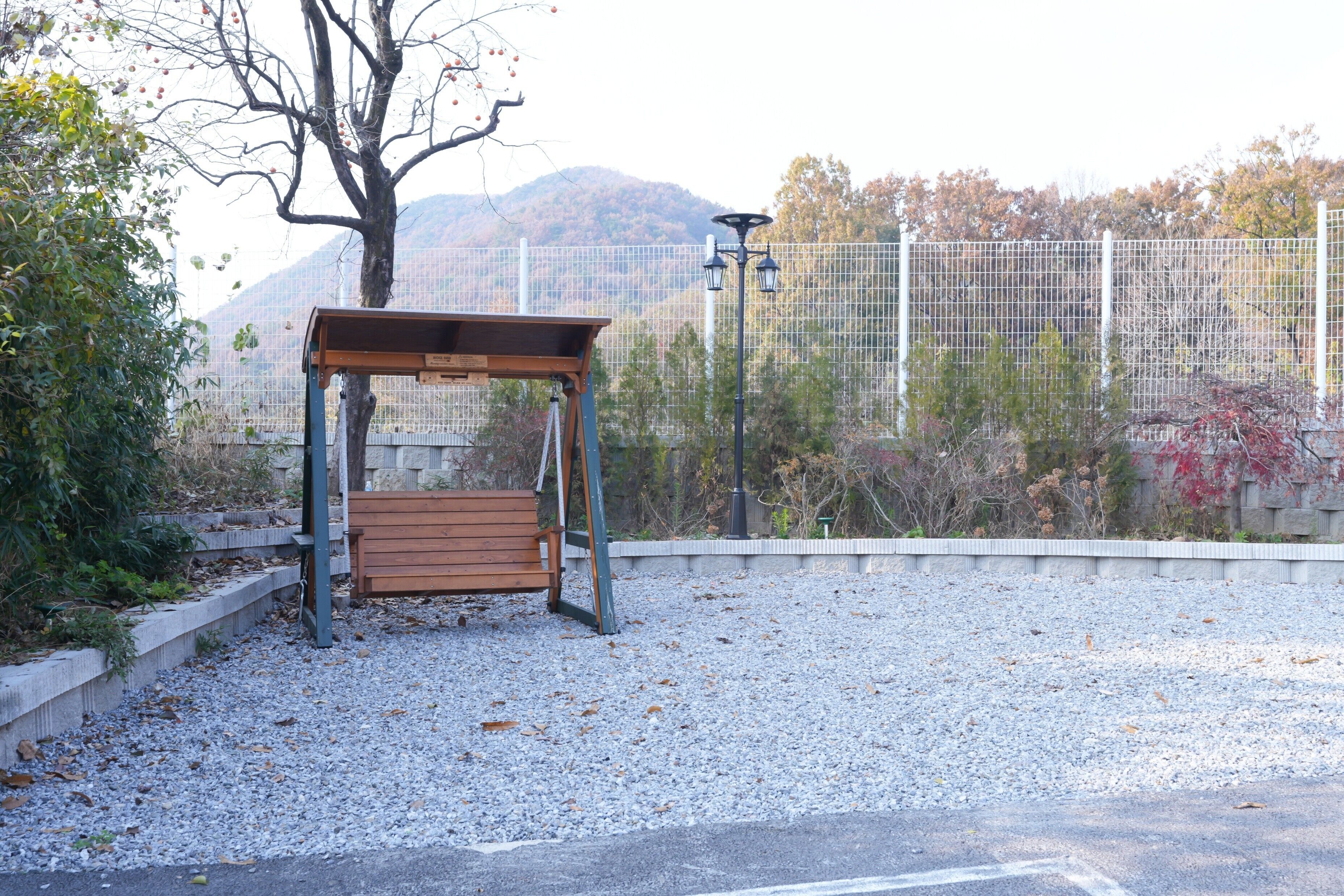 Un banco de madera bajo un refugio, con suelo de grava, rodeado de una cerca y árboles.