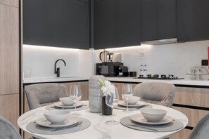 A modern kitchen with a dining area. It includes a round table with white plates, bowls, and wine glasses. There is a coffee machine and a kettle on the counter. - Modern Apartment with Rooftop Amenities (Dubai)