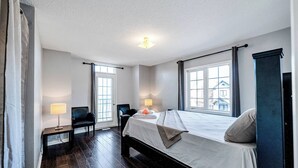 Habitación de hotel con una cama grande, dos lámparas de noche, una silla, una mesita y vistas al exterior a través de las ventanas.