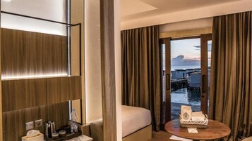 Habitación de hotel con una cama, una mesita de noche con una botella, un atril con artículos de aseo, una ventana con cortinas y vistas a un muelle y al agua.