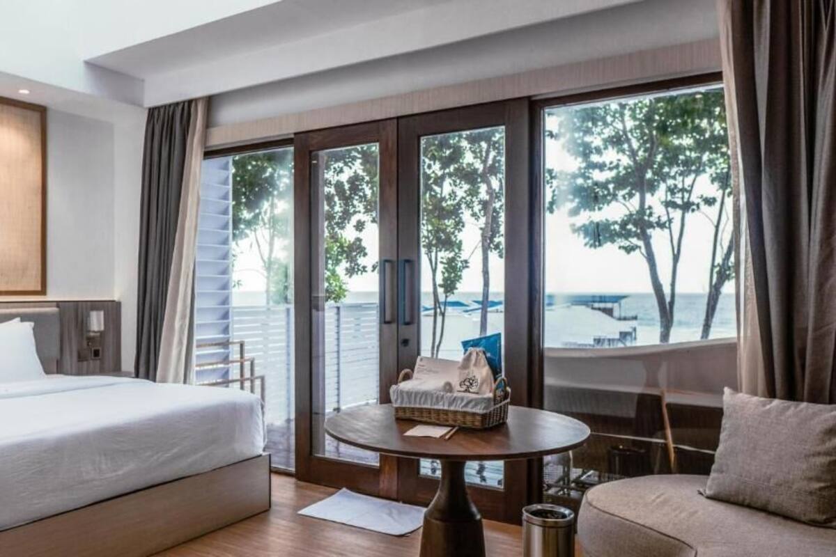 Habitación de hotel con cama, sofá, una mesita y vista al mar a través de amplios ventanales.