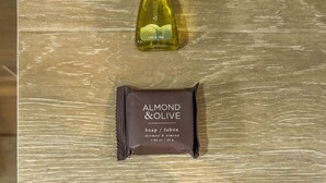 Superficie de madera con una botella de aceite de oliva, una pastilla de jabón de almendras y aceitunas, y una toalla blanca.