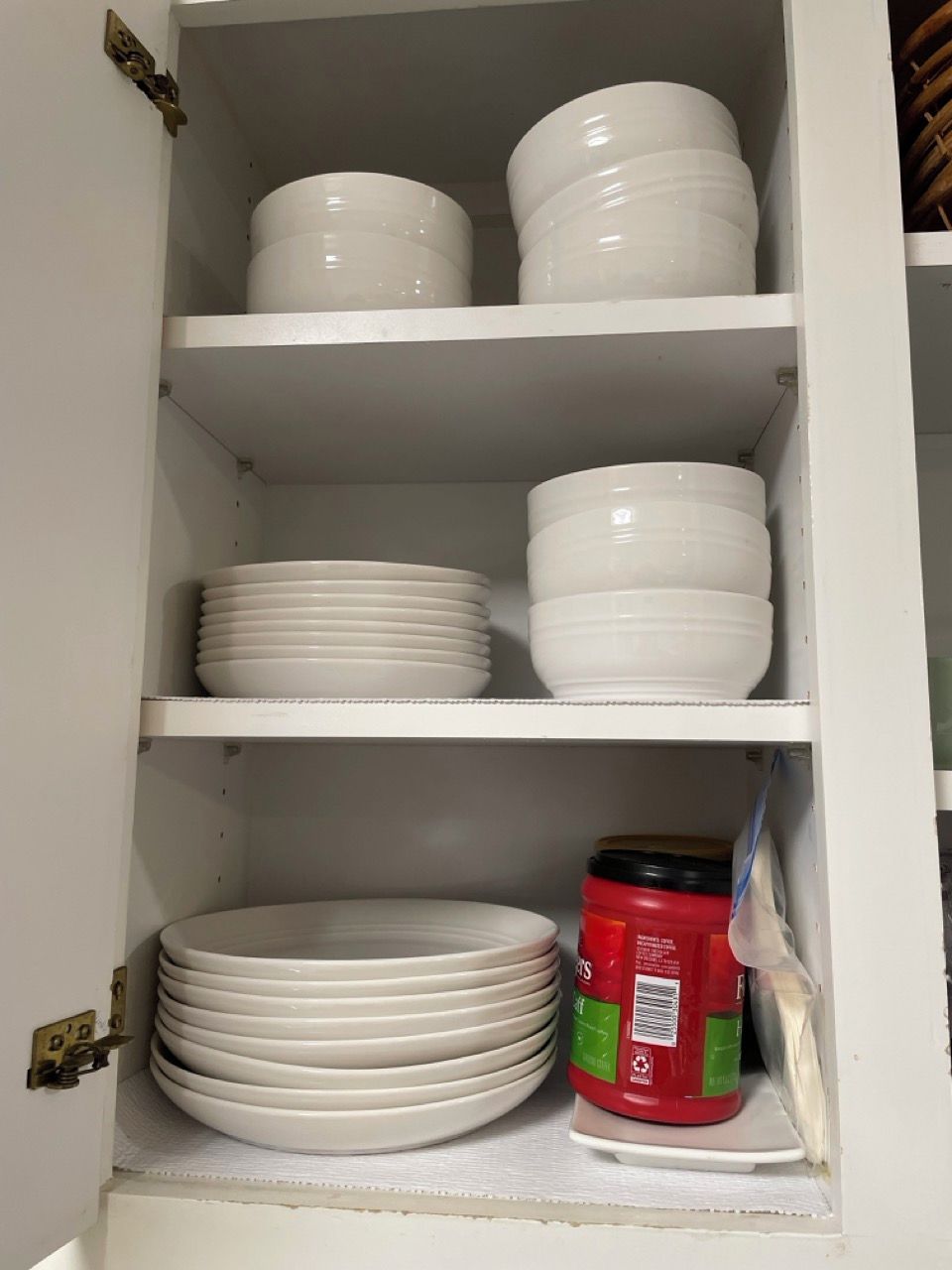 A kitchen cabinet with white storage shelves, containing stacked white plates and a red container with a green label.