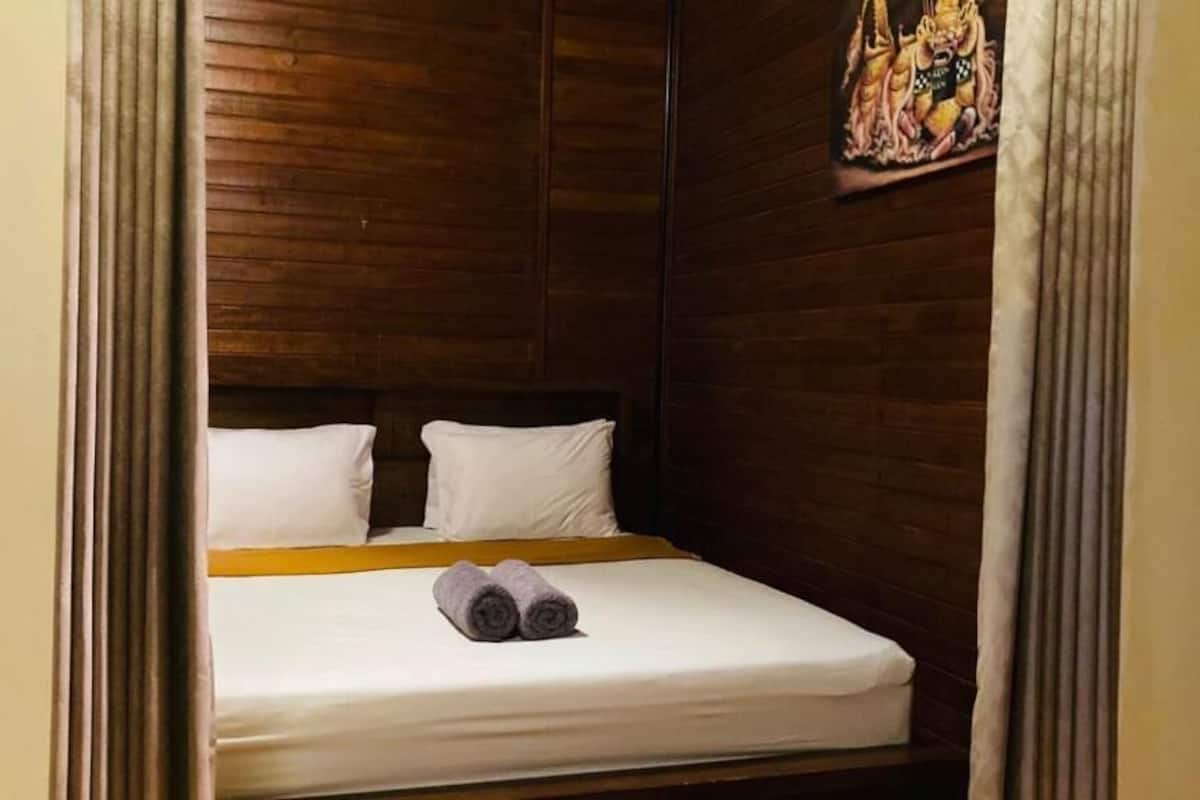 Una cama individual con ropa blanca y una toalla enrollada, paredes de paneles de madera y un adorno colgante.