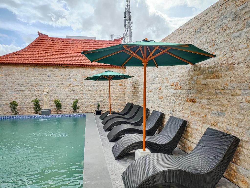 Area della piscina con lettini e ombrelloni, una parete in pietra e un edificio con tetto rosso sullo sfondo.