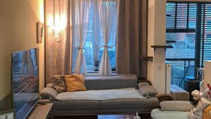 Een gezellige woonkamer met een bank, een bijzettafeltje en een televisie. Gordijnen, een raam en uitzicht op buiten.