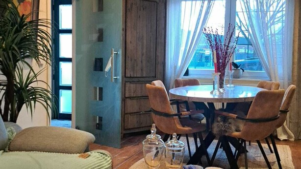Een gezellige woonkamer met een houten salontafel, een bank en een zithoek met een tafel en stoelen. Er is een raam met dunne gordijnen en uitzicht op het buitenleven.