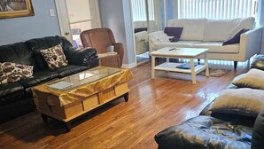 Ein Wohnzimmer mit einem schwarzen Ledersofa, einem weißen Sofa und einem Couchtisch mit Glasplatte. Es gibt einen Korbstuhl und einen weißen Sessel. Der Raum hat Holzböden und einen Deckenventilator.