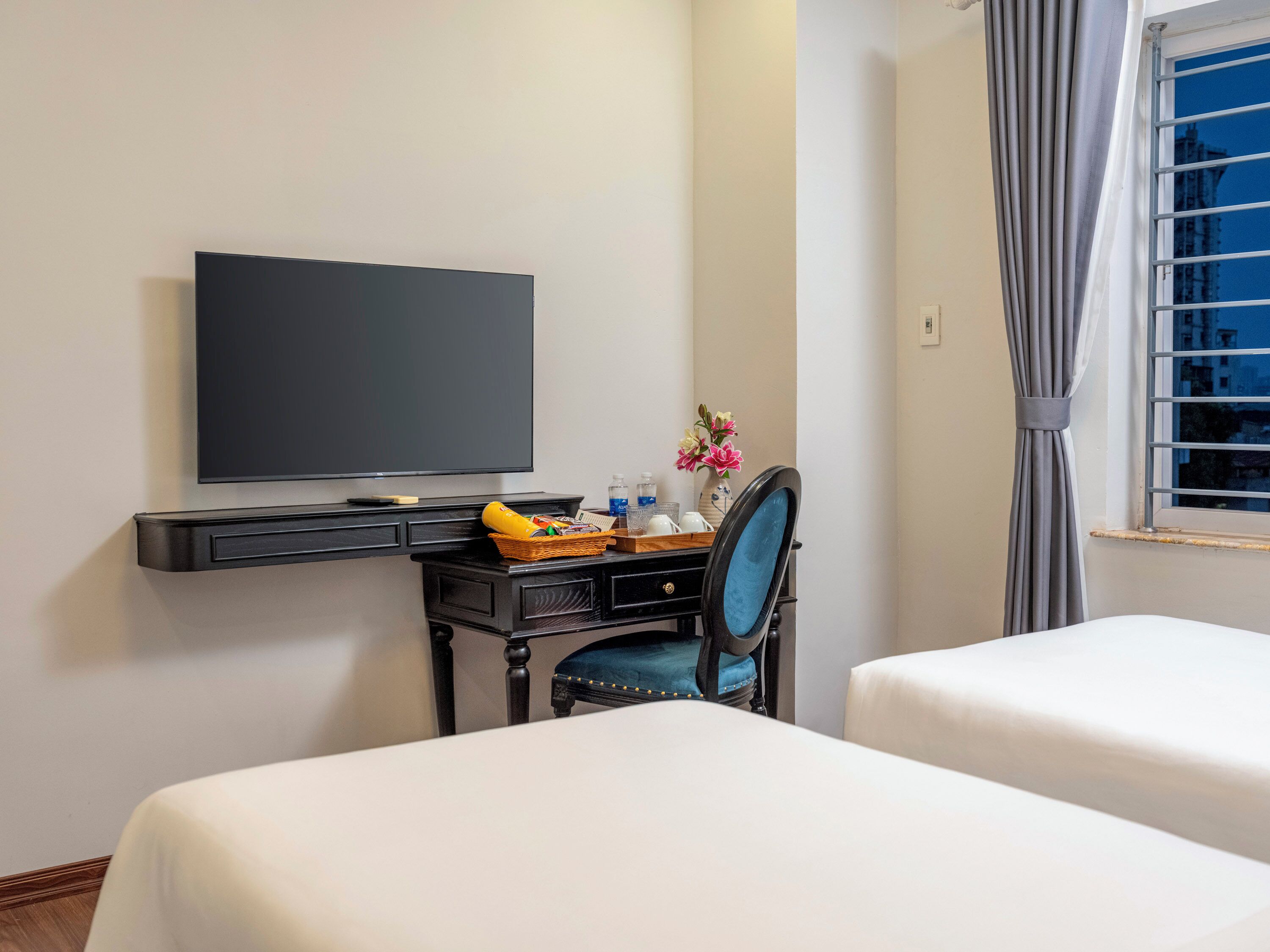 A hotel room with a flat-screen TV mounted on the wall, a desk with a basket of items, a chair, and two beds.