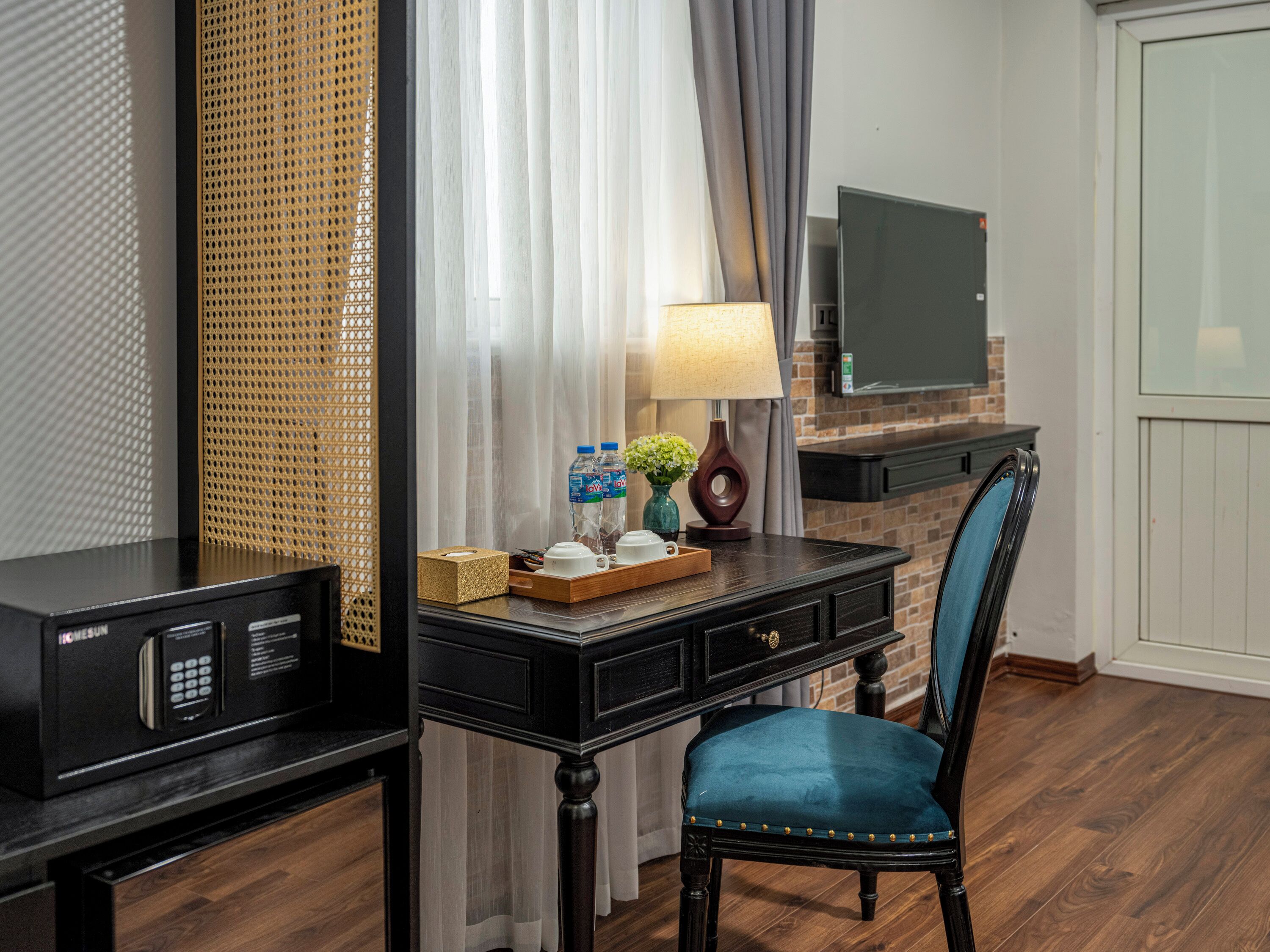 A hotel room with a desk, a chair, a television, a lamp, and a window with curtains.