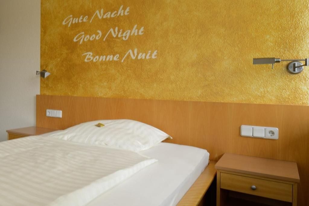 A hotel room with a bed, a nightstand, and a wall with a sign that says "Gute Nacht Good Night Bonne Nuit".