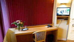 Ett litet rum med ett skrivbord i trä, en stol, en liten tv som är monterad på väggen och en dekorativ vas med blommor.