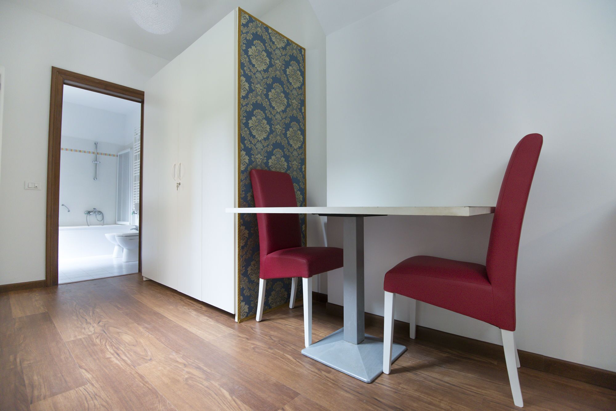 A room with a white table and two red chairs, a wooden floor, and a bathroom visible through an open door.