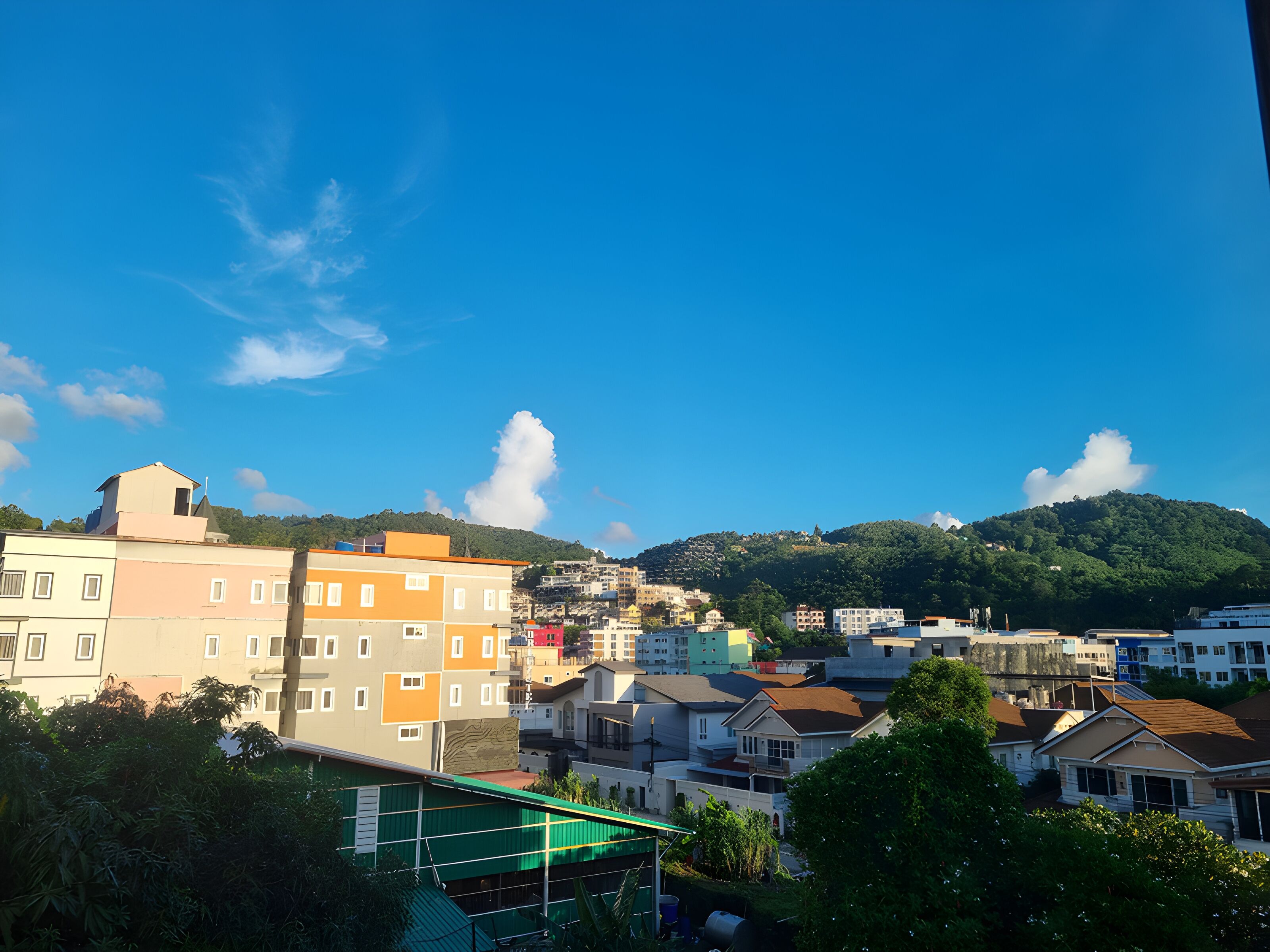 A residential area with multi-story buildings, a green hill in the background, and a clear blue sky.