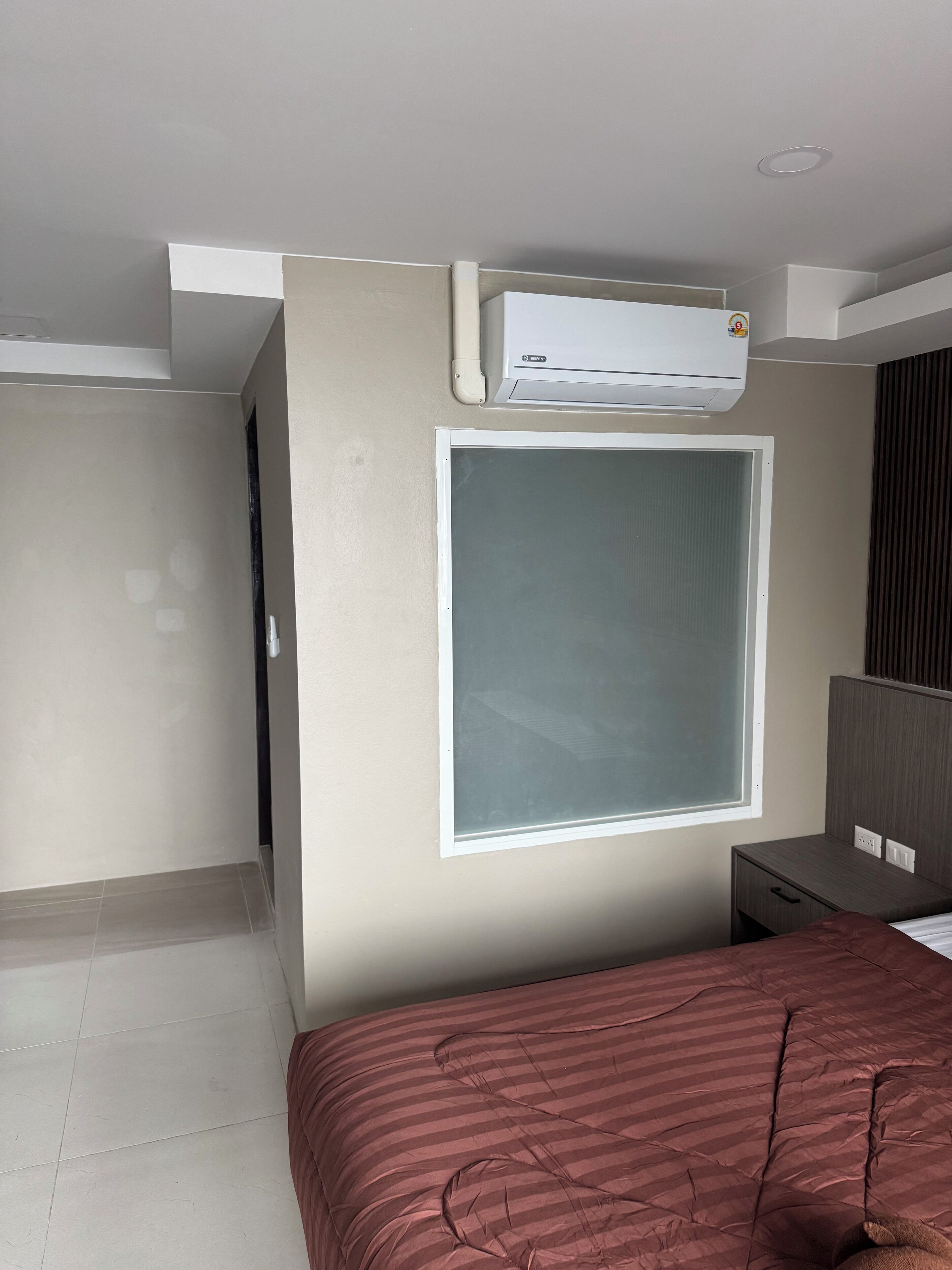 Una habitación de hotel con cama, ventana, equipo de aire acondicionado y una mesita de noche.
