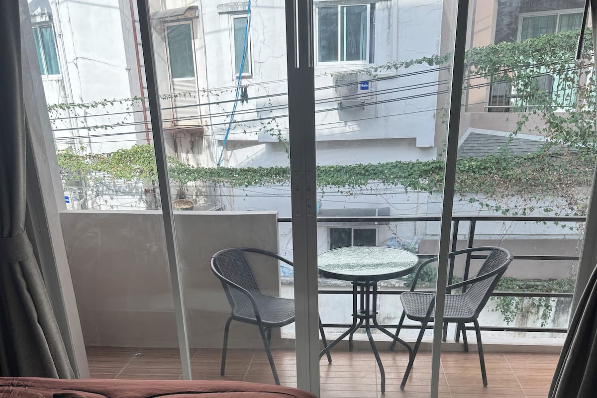 A hotel room with a large bed, a balcony with a table and chairs, and a view of the street and buildings outside.