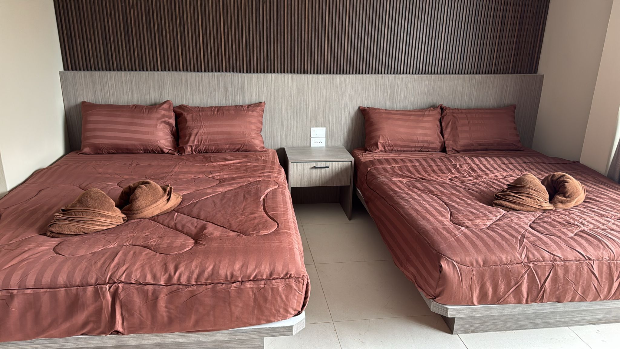 A hotel room with two beds, each with a matching headboard and pillows. There is a nightstand between the beds with a small drawer.