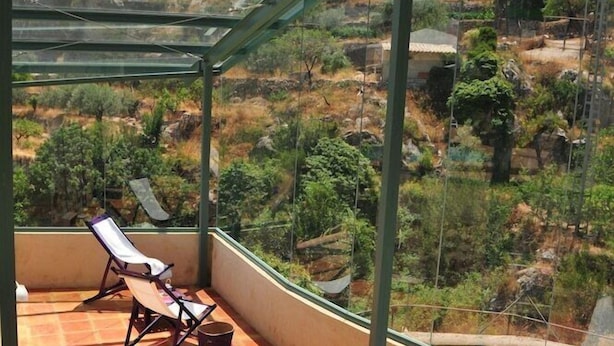 Een glazen balkon met uitzicht op een glooiend landschap, twee lounge stoelen en een tafeltje met spullen erop.