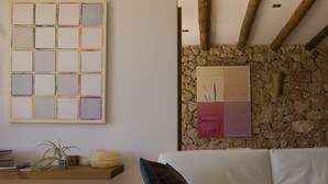 Een woonkamer met een witte bank, een houten plank met boeken en een plant, een stenen muur en twee abstracte schilderijen.