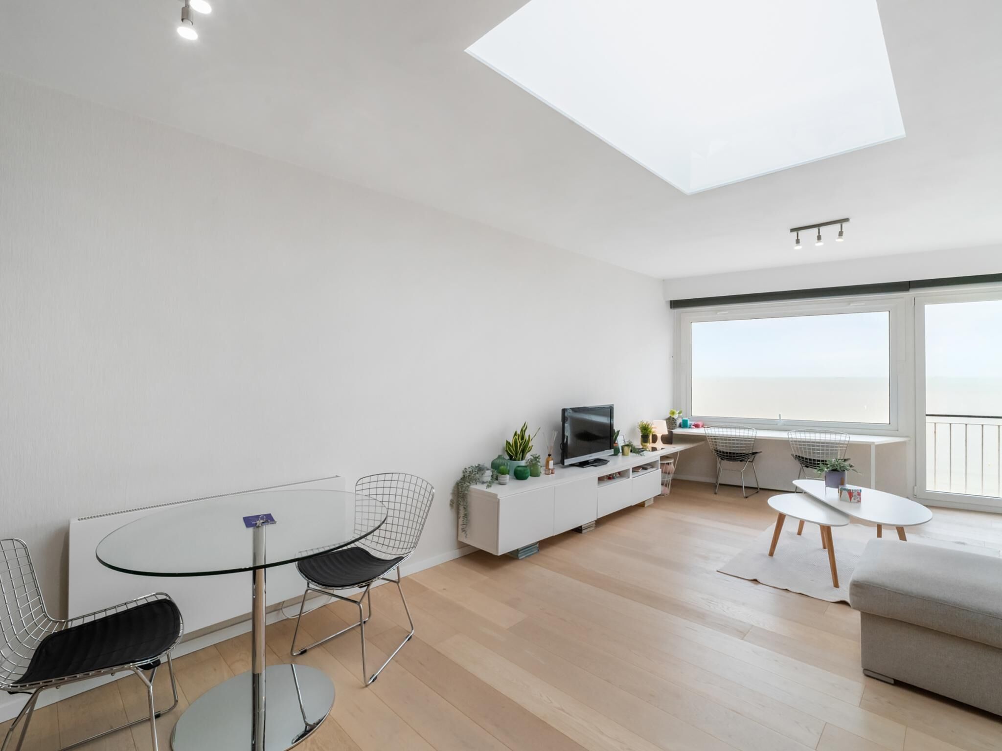 Ein modernes Wohnzimmer mit Glasdach, einem Essbereich mit Glastisch und Metallstühlen, einer weißen Unterhaltungsecke mit Fernseher und einem Balkon mit Blick.