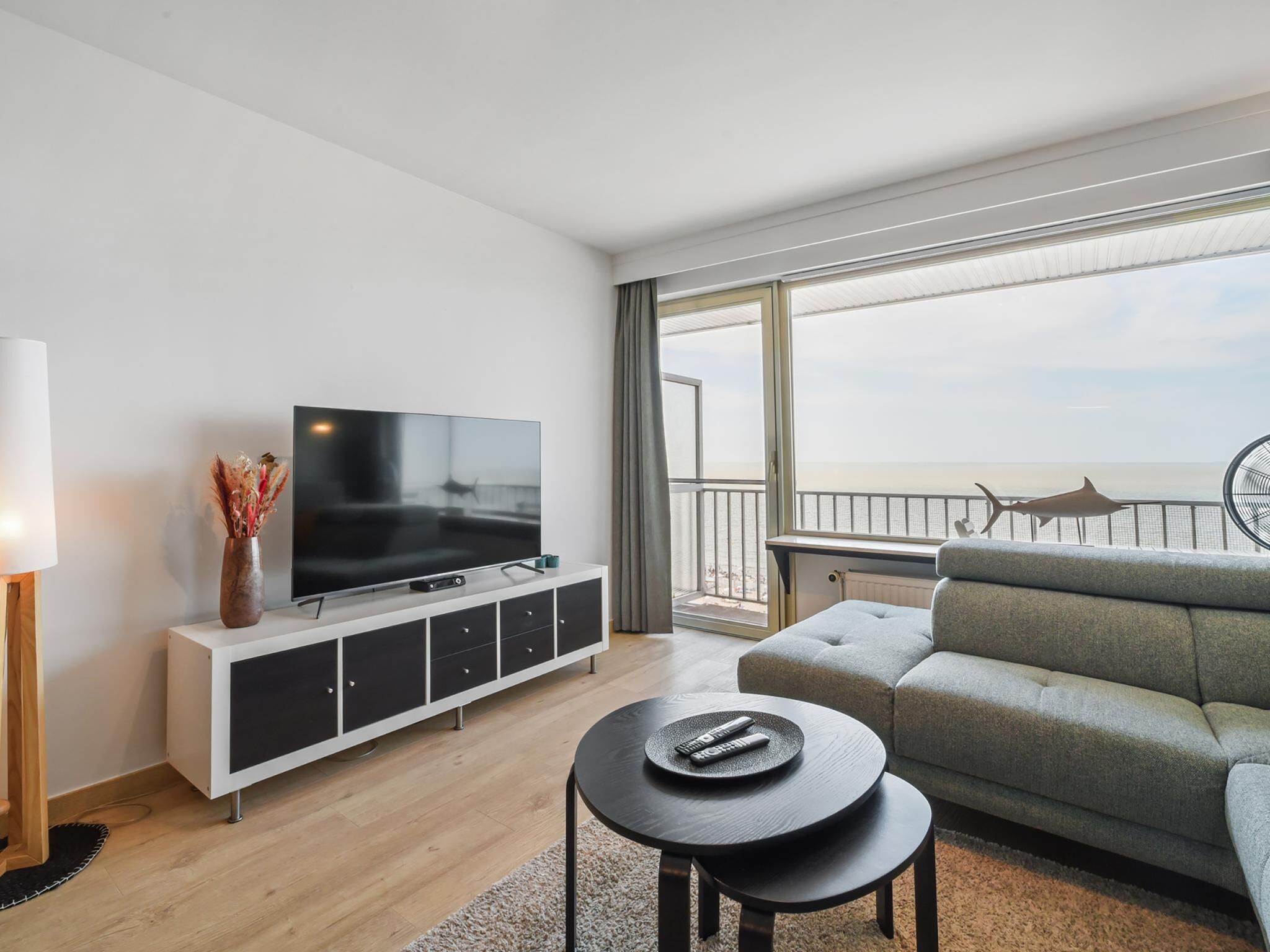 A modern living room with a flat-screen TV on a white cabinet, a grey sofa, a small round table with remotes, and a view of the sea through large windows.