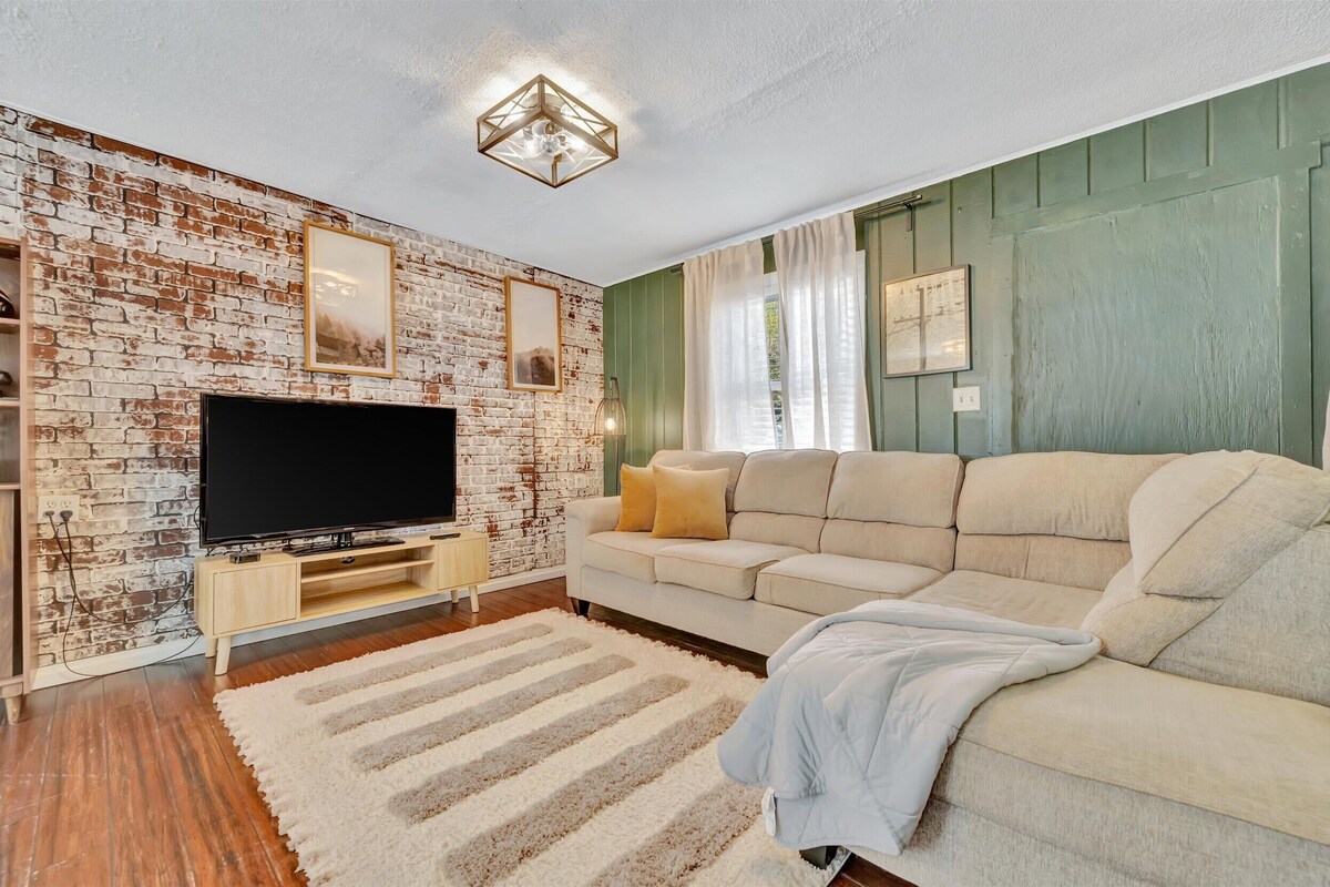Un salon avec un mur en briques, un téléviseur à écran plat, un canapé beige et un meuble de rangement en bois.