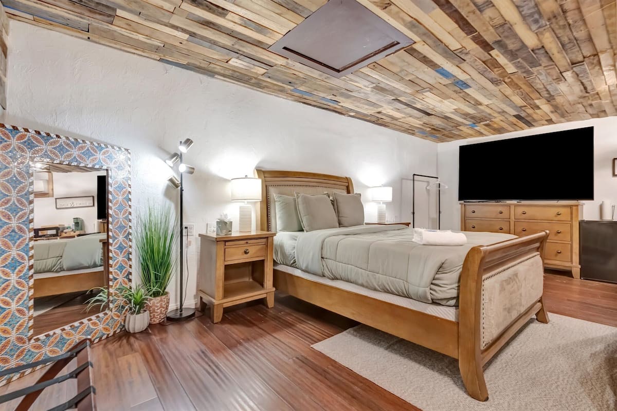 A bedroom with a wooden bed, bedside lamps, a large mirror, and a flat-screen TV.