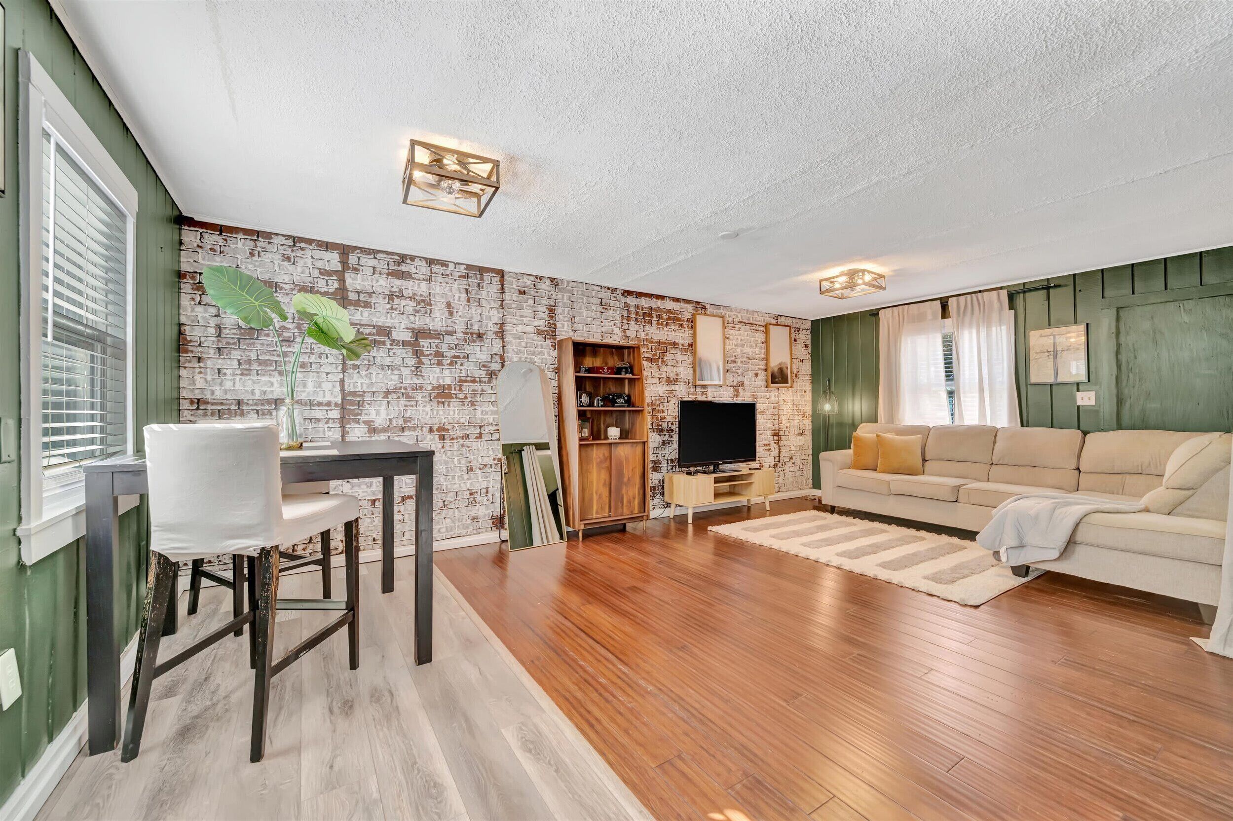 Ein Wohnzimmer mit Backsteinwand, Holzboden, einer Couch, einem Fernseher und einem Essbereich mit Tisch und Stühlen.