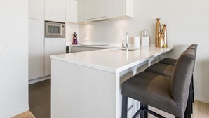 A modern kitchen with white cabinetry, a central island, bar stools, a built-in oven, and a coffee machine. - Beautiful apartment by the sea (Blankenberge)