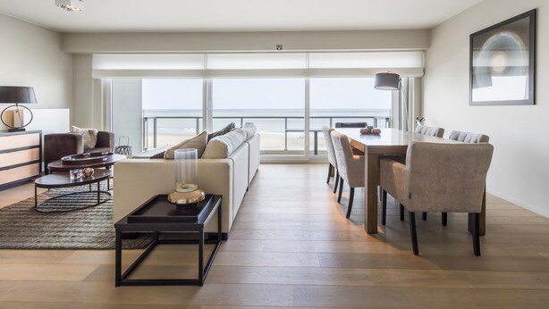A spacious living area with a large window overlooking the beach, a dining area with a table and chairs, and a seating area with a sofa and a coffee table. - Beautiful apartment by the sea (Blankenberge)